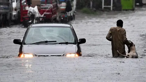 A man in a gray long-sleeved shirt inches along a flooded street with a white dog. He grabs its leash while a car wades through the same street during heavy monsoon rains in Rawalpindi, Pakistan on 17 July, 2025. 