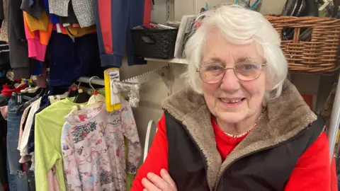An elderly woman with white hair and glasses smiles at the camera with her arms folded. She is wearing a red jumper and a dark gilet with a fur-lined collar, and stands inside a charity shop surrounded by rails of clothing. Children's nightwear and colourful garments hang on the rails behind her.

