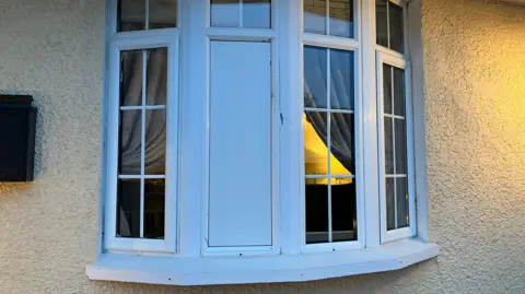 A white window frame with a number of sections of glass panes. A large section of the window is covered with a white plank of wood. 