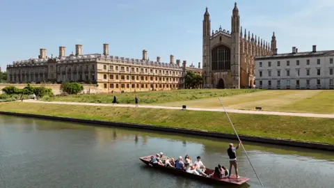 A view over the River Camb towards King's College Chapel and other ornate college buildings. A punt with around a dozen sightseers on board is seen in the foreground, being pushed along the river by a punting chauffeur, who is wearing a white T-shirt and waistcoat. He is holding a long metal staff, which is used to push against the bottom of the river and propel the boat along. Beyond the river are lawns leading up to the buildings in the middle distance. The sky is blue and it appears to be a gloriously sunny day.