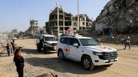 Reuters A Palestinian girl watches two Red Cross team vehicles assisting in the search for the last dead Israeli hostages drive through Gaza City (8 December 2025)