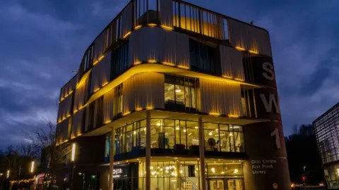 A modern five tiered building with a glass frontage is illuminated in yellow lighting at night. 