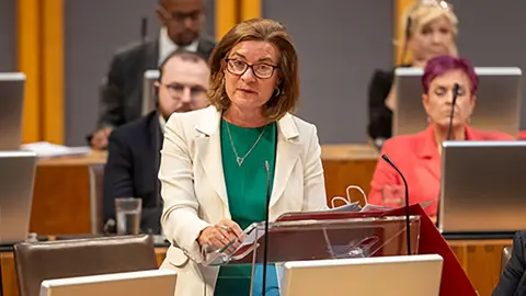 PA Media Eluned Morgan stood addressing the Senedd, in a white jacket and green dress.