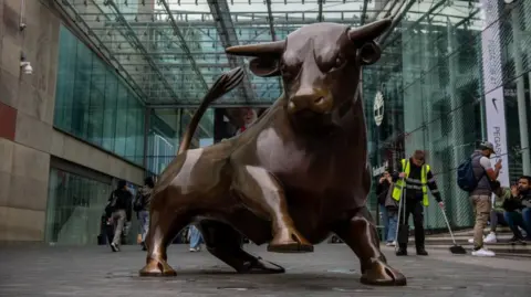 Getty Images A large bronze statue of a bull outside a shopping centre, which has a glazed front. The bull has one leg raised and an angry expression, as if it is about to charge. Pedestrians and a street cleaner in a fluorescent jacket can be seen behind it.