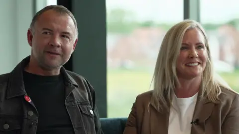 A man sits on a sofa alongside a woman. There is a large window behind them. Both are looking away from the camera. The man has short hair which is slightly grey. He is wearing a dark grey denim jacket with a black T-shirt underneath. The woman has long blonde hair and wears a light brown jacket with a white T-shirt underneath. 