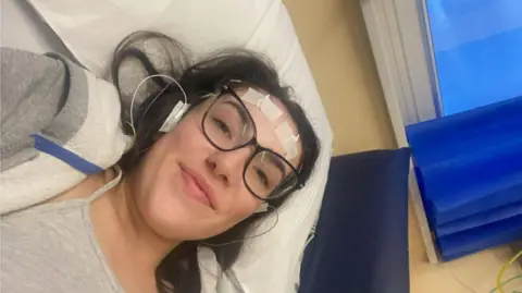 Sophie Morgan Sophie is led down in a hospital bed. She wears black square rimmed glasses and has various wires taped to her forehead. She has brown eyes and brown hair. She wears a grey vest and a grey zip up hoodie. 