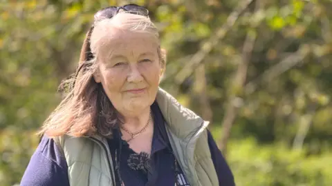 A woman looking at the camera, she has light brown and blonde hair with black sunglasses on top off her head. She is wearing a grey/green gilet and a navy polo shirt.