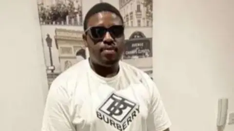 A man stands with a white T-shirt that reads "Burberry" across the front. He is wearing black, square sunglasses and standing in front of a white wall with a black and white picture on it.