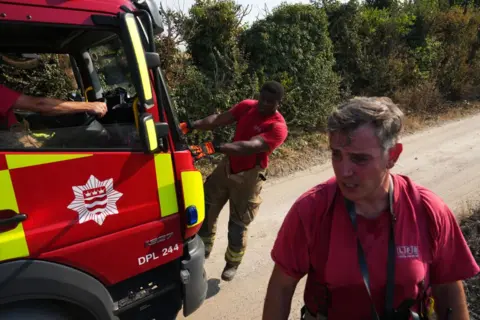 Getty Images Firefighters and engine attending a grass fire in summer 2022, Firefighters wearing red T-shirts and yellow protective trousers look weary and sooty.
