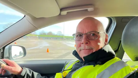 Man with moustache and glasses and hi-vis vest behind steering wheel, looking at camera.