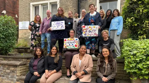 A group of people pose together outside a brick building, with some standing and others sitting on steps, holding colourful hand‑made signs with words and drawings.
