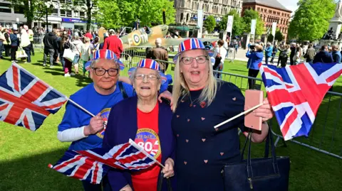 three women festooned in Union flag colours wave flags, wearing T shirts comemmorating VE Day. They are standing outside Belfast City Hall
