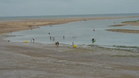 Qays Najm/BBC Part of the sandy beach at Wells-next-the-Sea in Norfolk. People can be seen in the water at the beach. The sky is blue.
