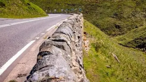 Sheffield City Council A winding road with a sharp bend on a hillside has a low stone wall separating it from a deep drop into the countryside