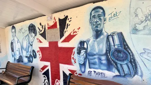 John Devine/BBC Graffiti art work inside a bus shelter shows three boxers from Chatteris, from left to right, Dave 'Boy' Green, Eric Boon and Jordan Gill. Two wooden benches are in front of the artwork which is done in black and white, with part of a Union flag in the middle.