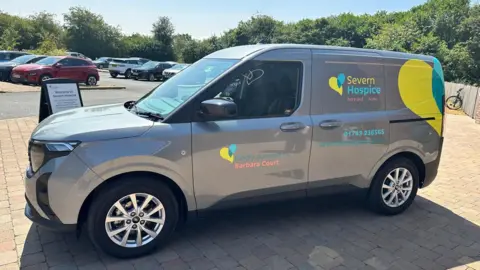 A small grey van with yellow and blue lettering and logos on it. Writing says "Severn Hospice". On the passenger door it reads "Kindly donated by Barbara Court".