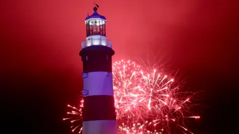 Plymouth City Council Red-and-white striped Plymouth lighthouse stands in the foreground as vivid red fireworks burst behind it, lighting up the night sky and surrounding smoke. Spectators gather along the waterfront below the display.