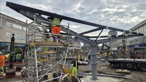 Mark Lowry The image shows a busy construction site in a city centre. A large steel structure is being assembled in the middle of the scene. The structure has a thick central column and long metal beams stretching out from the top, suggesting it is the early stage of installing a canopy or similar overhead feature.