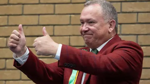 Getty Images Plaid Cymru's newly elected Caerphilly Member of the Senedd Lindsay Whittle at the count. He is wearing a maroon suit jacket with blue check marks and a green tie with a white shirt. He is giving a double thumbs up and is smiling. Lindsay is a middle aged man with short grey hair