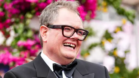 Comedian Alan Carr in a bow tie and black suit against a blurred background of flowers
