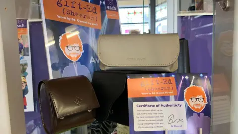 East Anglia's Children's Hospices Items including a grey leather glasses case are on display in a EACH charity shop in Framlingham. The items are next to a purple poster with a graphic of Ed Sheeran on and a purple certificate of authority.