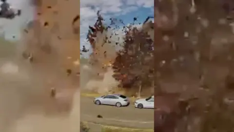 Debris flying through the air mid-explosion, on a tree-lined street with parked cars.