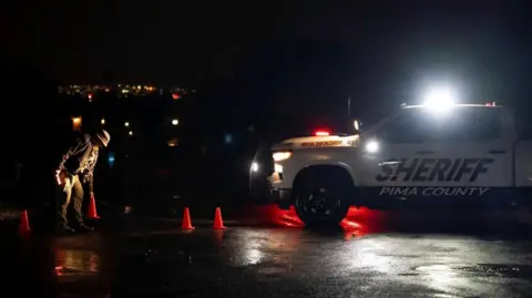 Reuters An official examines the ground near orange traffic cones at night, illuminated by headlights from a Pima County Sheriff’s vehicle.