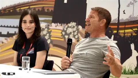 John Fairhall/BBC Amy Meek on the left sitting down next to Steve Backshall. She is wearing a black T-shirt and has short dark hair. Steve Backshall is also sitting down and he is wearing a grey T-shirt and is holding a microphone.