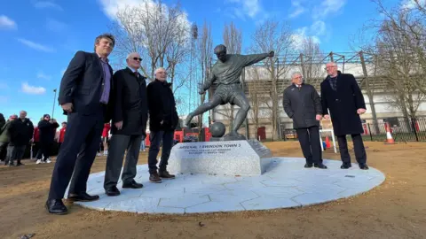 A group of men stand around a new statue of Swindon Town player Don Rogers. The statue shows him about to kick a ball. In the background more people and the County Ground Stadium can be seen