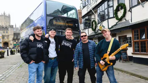 First Bus 3SEVEN6 members are standing next to each other in front of a bus. They are by a pub in Glastonbury. One of them s holding a guitar.