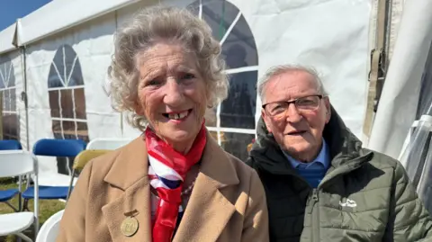 A lady is pictured on the left of the image wearing a brown coat and a red and white scarf. She has grey hair and is smiling at the camera. The man on the right is wearing a green jacket and glasses. He has grey hair.