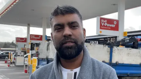 A man with short black and grey hair standing on a petrol forecourt. He is wearing a white T shirt and grey coat and looking down the camera.