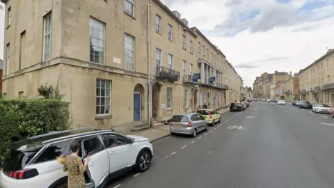 Google Maps A view of Beaumont Street . There are cars parked on the sides. Someone is seen getting in to a car, holding a bouquet of flowers. It is a cloudy day.