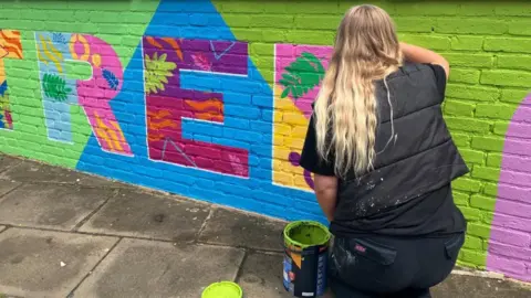 BBC/Hannah Sackville-Bryant A closer-up view of a vibrant mural, painted along a low brick wall along a riverside path. The mural depicts the word 'Rowntree' painted in bright multicolours against predominately lime-green background. Each letter also contains leaf and flower motifs within it. In the corner, a woman with long blonde hair and wearing black paints onto the mural.