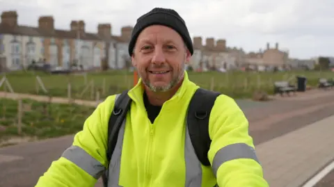 Shaun Whitmore/BBC Robbie Kenny smiles at the camera as he sits on a bicycle on a road. A row of houses can be seen in the distance behind him. He wears a black beanie hat and a hi-vis jumper. He has a rucksack on.