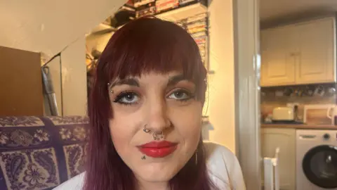 A young woman with red hair, facial piercings and red lipstick she is sat on a sofa in a property.