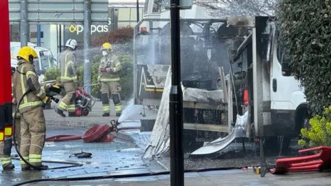 Three firefighters can be seen wearing their kit, which is beige with high vis yellow detailing and yellow protective helmets. One firefighter holds a hose and it washing down the truck, while two others in the background hold another piece of equipment. The truck is scorched black.