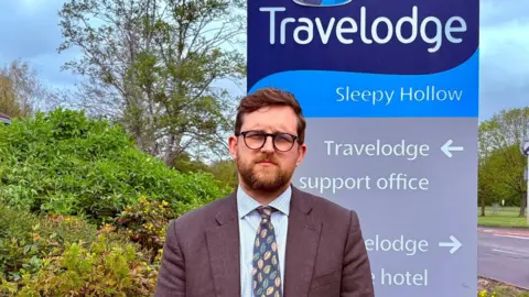 Freddie Van Mierlo Freddie van Mierlo stands in the middle of the image wearing a blue shirt with a navy tie with leaves on it. He is wearing a light brown suit jacket. He has a brown beard and brown hair and is wearing black rimmed glasses. Behind him, in the Travelodge brand colours of navy and white, is a sign for the Travelodge HQ.