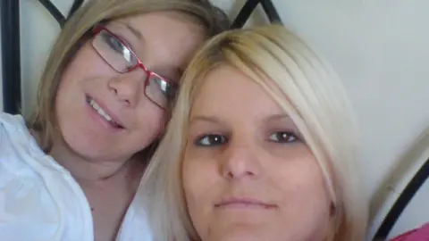 Bethan Callaghan (right) picture with her sister Chloe Callaghan. Chloe has dark blonde hair with a side fringe and wears pink rectangle rimmed glasses. She has blue eyes and smiles at the camera. Bethan has brown eyes and bright blonde hair. She looks directly at the lens and smiles. 