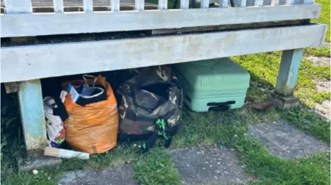 Devon and Cornwall Police Bags and a green suitcase are stored under a raised wooden deck on grass and paving.
