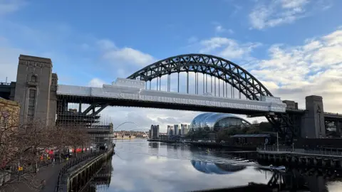 The Tyne Bridge. Large white sheets cover much of the bridge where repair work is being carried out. Scaffolding can be seen underneath the bridge by the concrete towers.