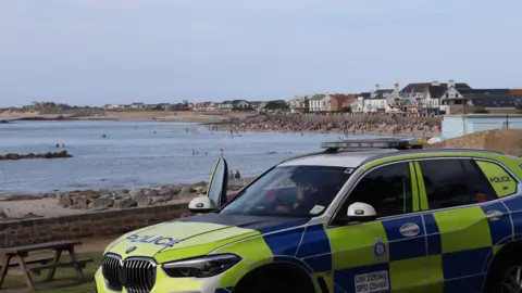 A Guernsey Police car in the usual green and blue police branding parked up on the road over looking a beach with lots of people sat on it and swimming in the sea. There is an officer sat in the car looking out at the people with the car door open. 