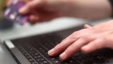 A left hand types into a laptop keyboard, with the right hand in soft focus holding a bank card.