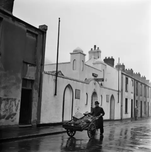 Bert Hardy/Getty Images A fish seller pushes his barrow past a mosque. He is wearing a fedora-style hat.