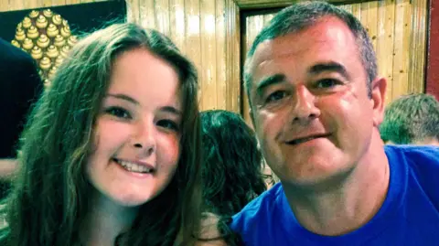 A family photo of Emily and her father David. They are sitting close together indoors, smiling and posing for a photo. Emily, on the left, has long brown hair, and David, on the right has short greyish hair and is wearing a blue shirt. Behind them is wooden panelling.
