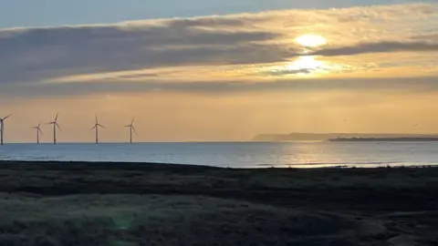 Weather Watchers/Superted A warm yellow and blue sky with horizontal clouds stretching across it. There are wind turbines and cliffs on the horizon and in the foreground there is a grassy cliff edge.