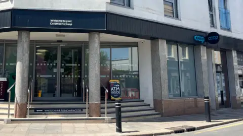 BBC A wide shot of the co-operative. It is fronted by two grey pillars, and has a dark coloured sign.