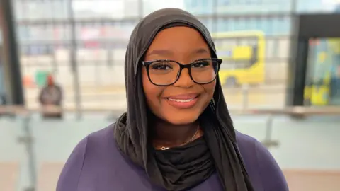 BBC A smiling, young woman wearing black glasses and a hijab. 