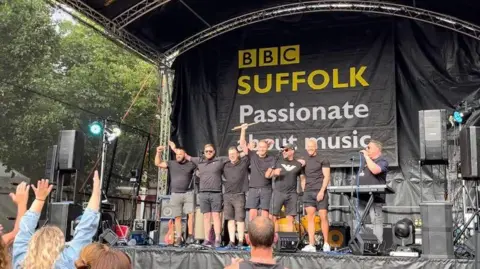 The 2023 Ipswich Music Day event. A group of male performers stand on a stage looking out at the crowd and smiling. Crowds in front of the stage wave their hands in the air. A banner on the stage reads: "BBC Suffolk, Passionate about music."