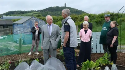 PA Media King Charles III standing in an allotment. There is a group of men and women standing behind him and he is talking to a man. 